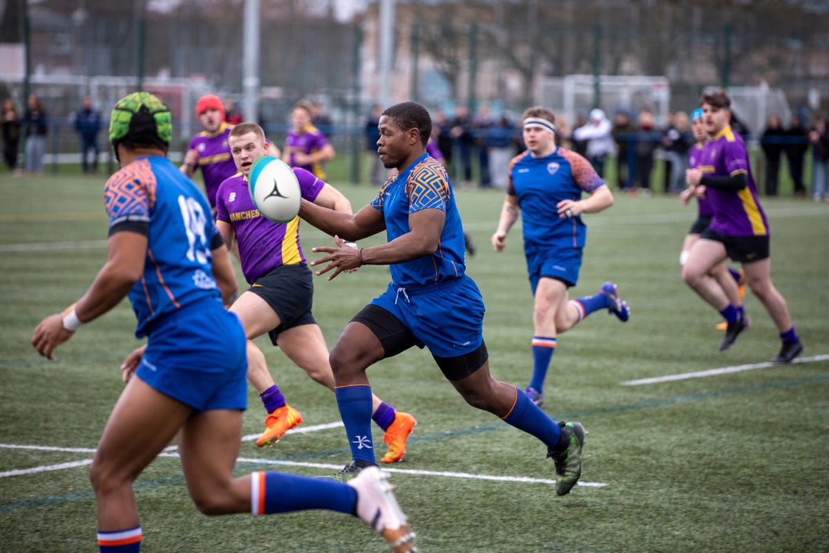 A dynamic rugby match scene on a green turf field, featuring players in blue and purple jerseys running and tackling. A player in a blue jersey is captured mid-run with a rugby ball, surrounded by teammates and opponents.