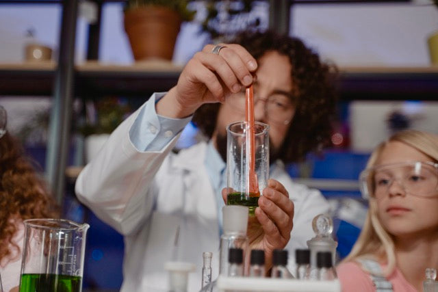 A teacher in a white lab coat holds a glass test tube with green liquid and an orange stir rod in a laboratory setting, with another person in safety goggles partially visible in the foreground and background.
